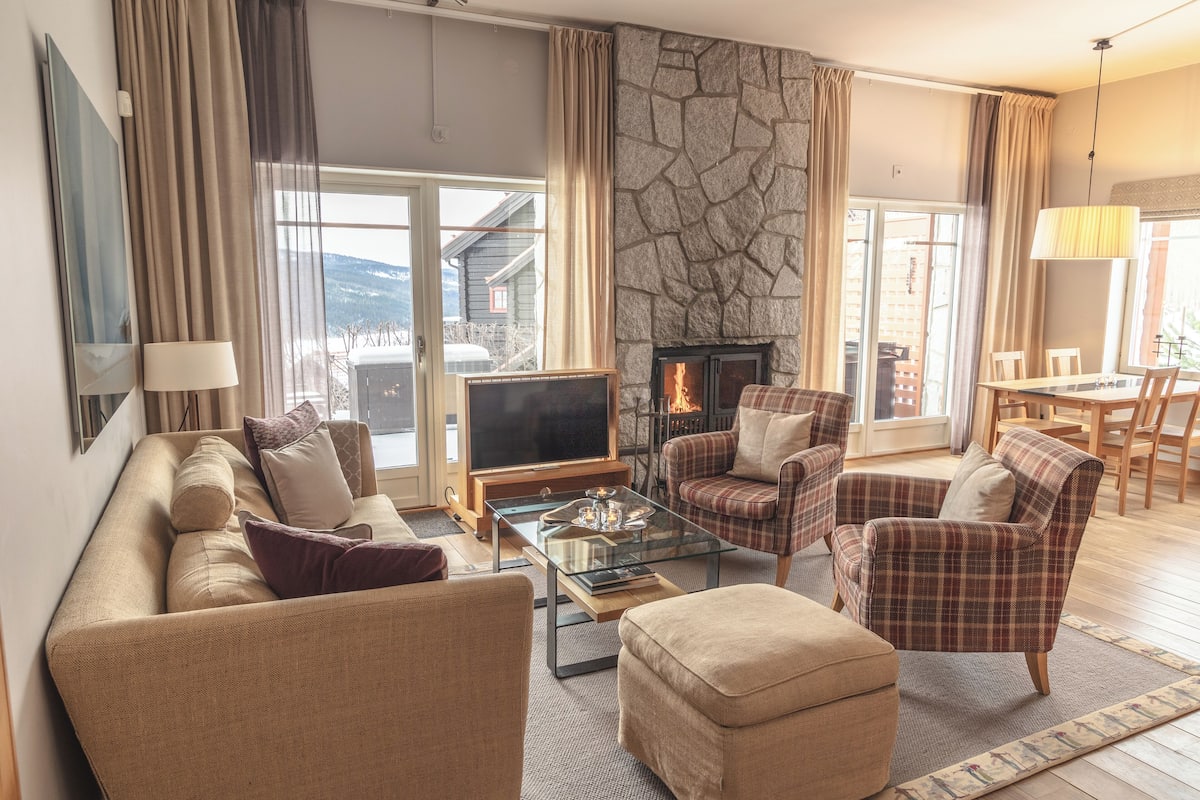 Un salon chaleureux avec une cheminée en pierre, des sièges confortables et une grande fenêtre donnant sur le paysage extérieur.