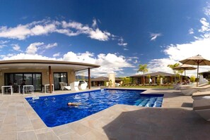 A swimming pool with a person swimming, surrounded by lounge chairs and umbrellas, with a covered seating area and a clear sky. - Eden Appartel Koné (Kone)