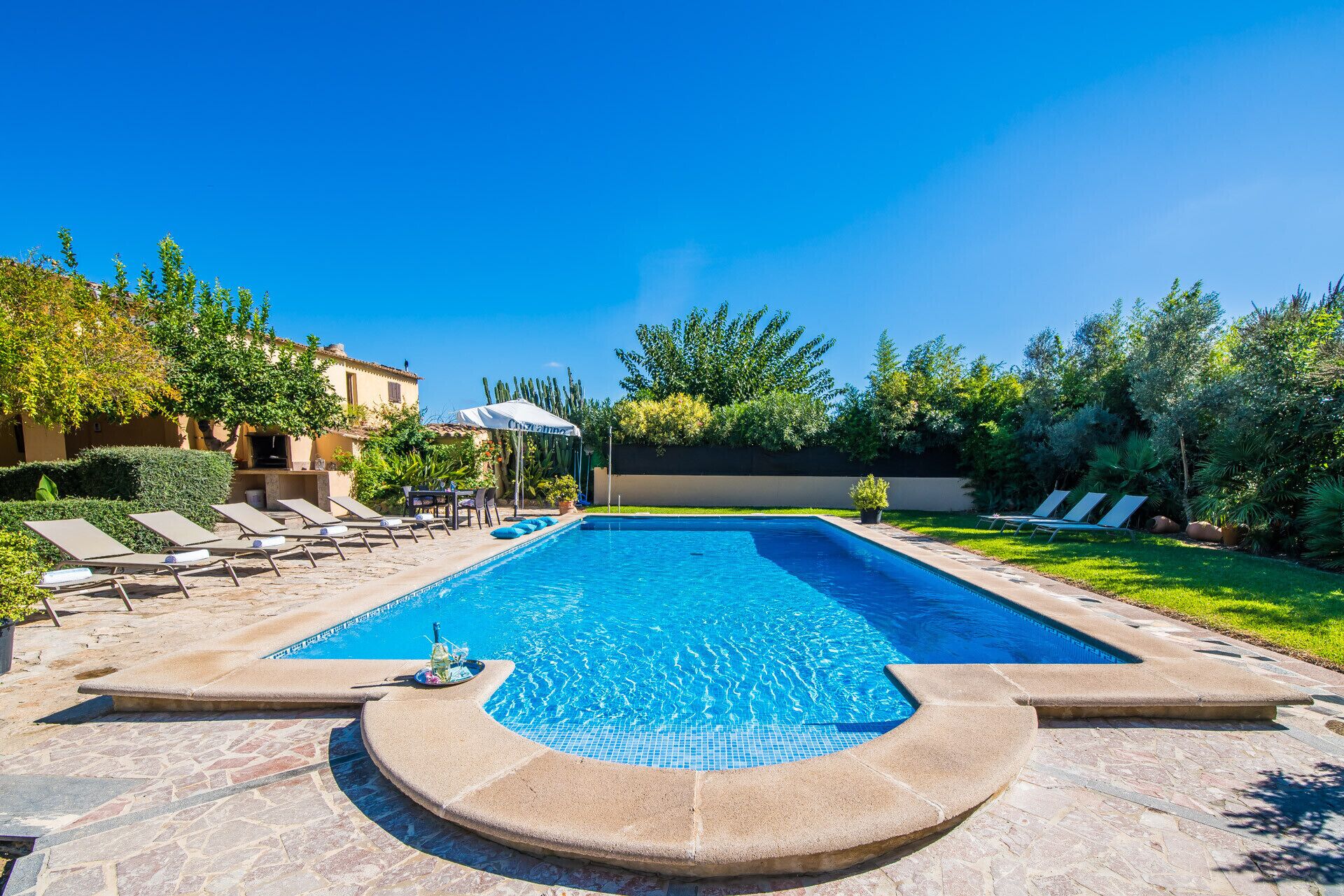 Una piscina rectangular con borde de azulejos, rodeada de sillas de descanso y una pequeña mesa con sombrillas.