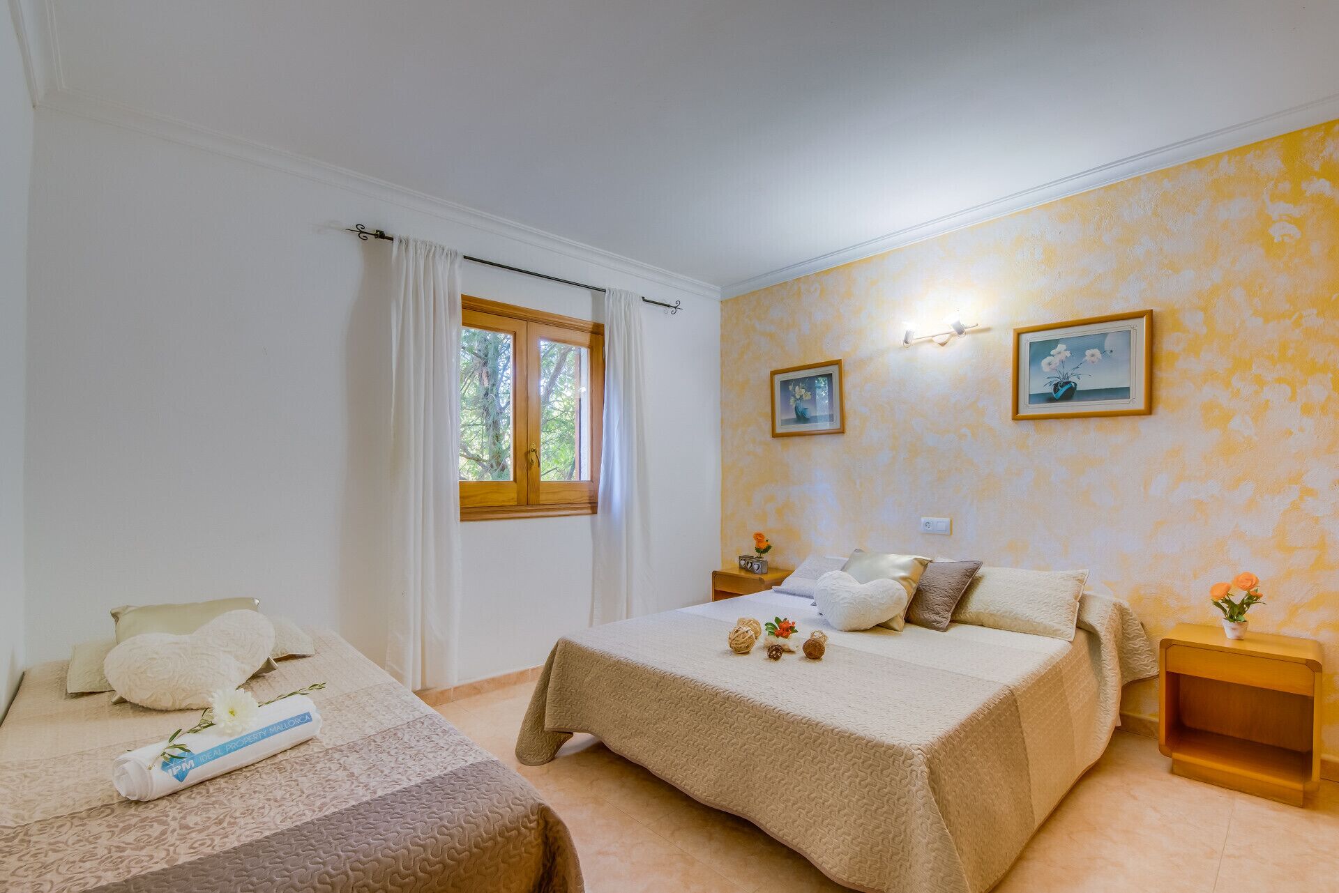 A bedroom with two beds, a window with curtains, a bedside table with a clock, and two framed pictures on the wall.