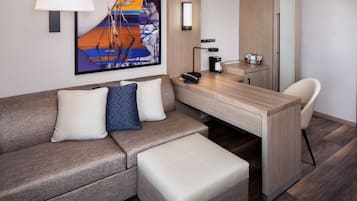 A modern hotel room with a sofa, a small table, a chair, a desk, and a refrigerator.
