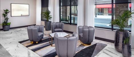 A modern hotel lobby with a marble floor, grey upholstered chairs, a small table, and large windows with curtains.