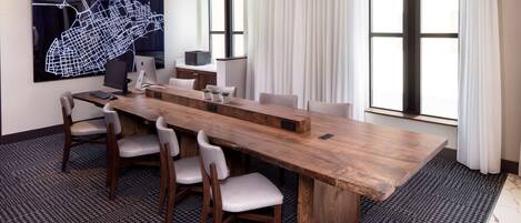 A conference room with a long wooden table, six chairs, a large abstract wall art, a desk with a computer, and large windows with curtains.