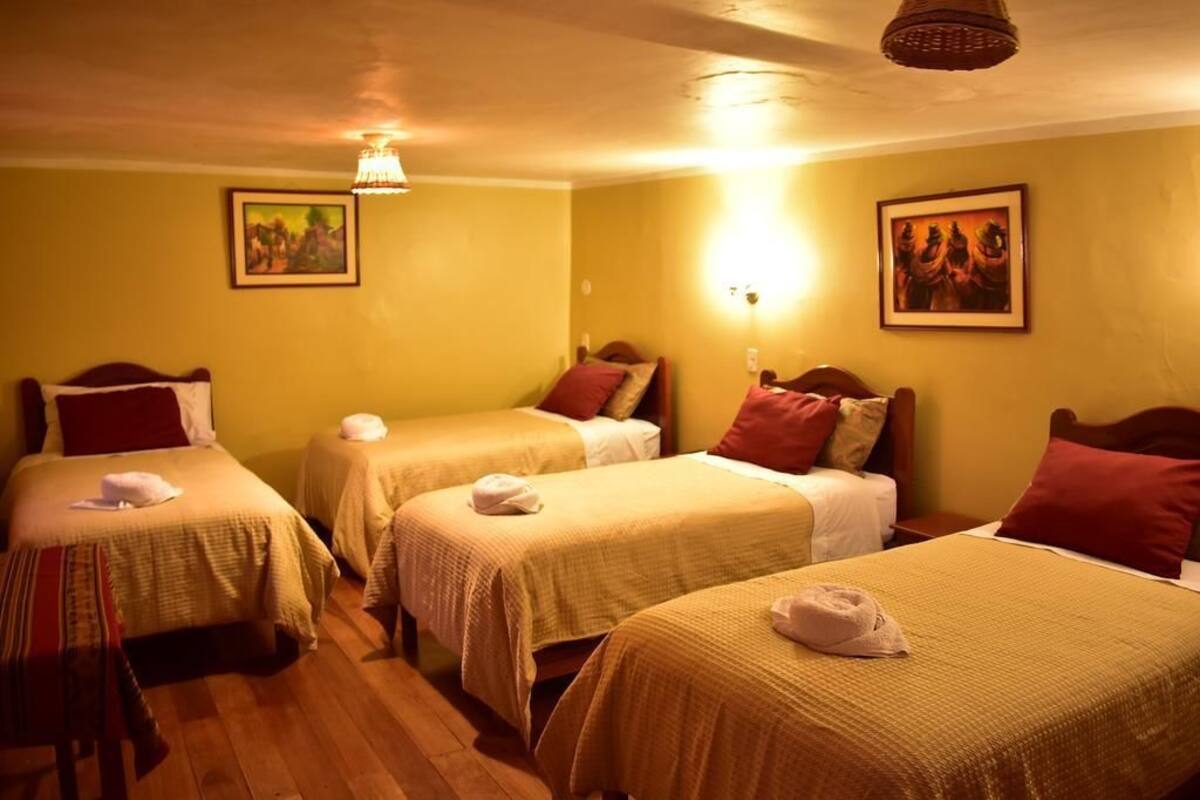 Habitación de hotel con tres camas individuales, cada una con una almohada roja y una toalla blanca. Las paredes son amarillas y el suelo es de madera.