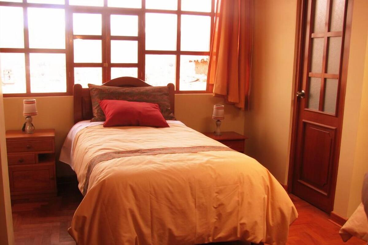Un dormitorio con cama, mesitas de noche de madera, una ventana con cortinas y una puerta.