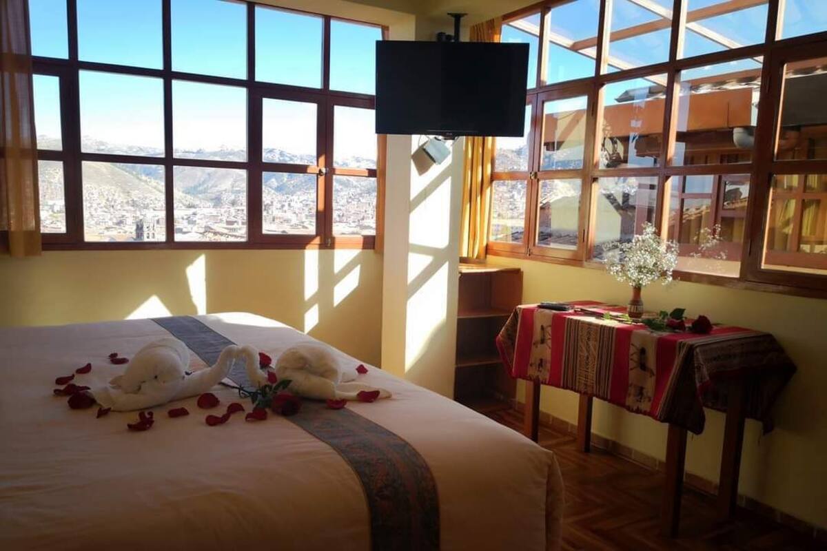 Habitación de hotel con una cama grande, un televisor, una mesa con un arreglo floral y vistas a un paisaje urbano a través de amplios ventanales.