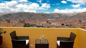 A balcony with wicker chairs and a small table, overlooking a town with red-roofed buildings and mountains in the background. - Kuska Guest House (Cusco)