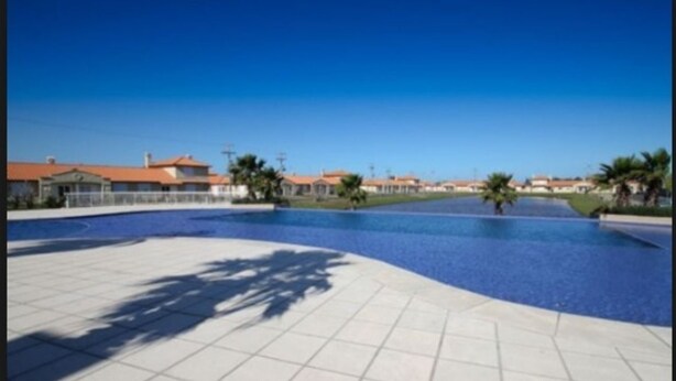 A swimming pool area with tiled flooring, palm trees, and residential buildings in the background.