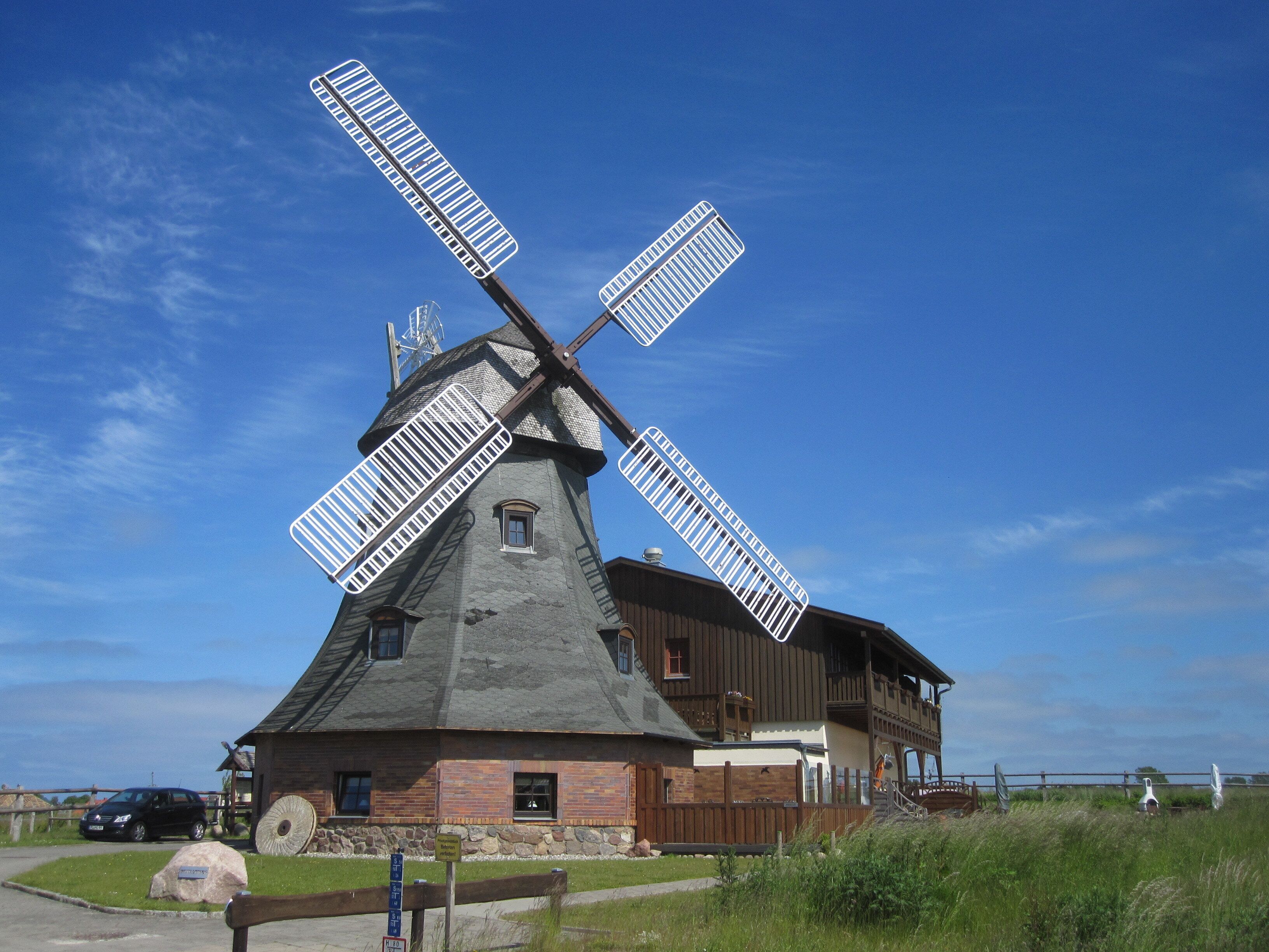 A traditional windmill with a wooden structure and a stone base, surrounded by a grassy area and a wooden fence.