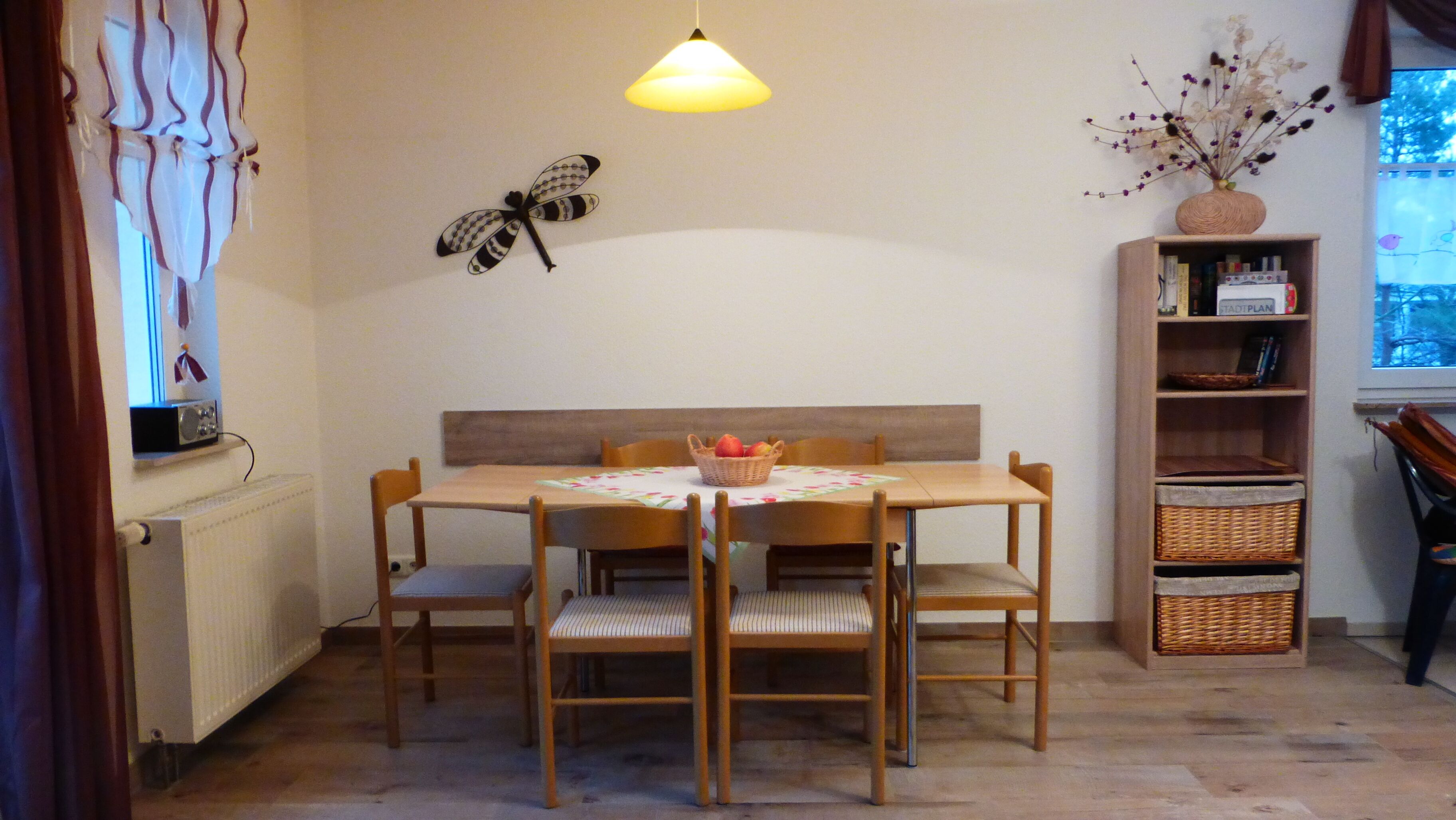 Een eetruimte met een houten tafel, vier stoelen, een hanglamp, een wanddecoratie van een vlinder, een mand met fruit en een boekenkast met diverse spullen.