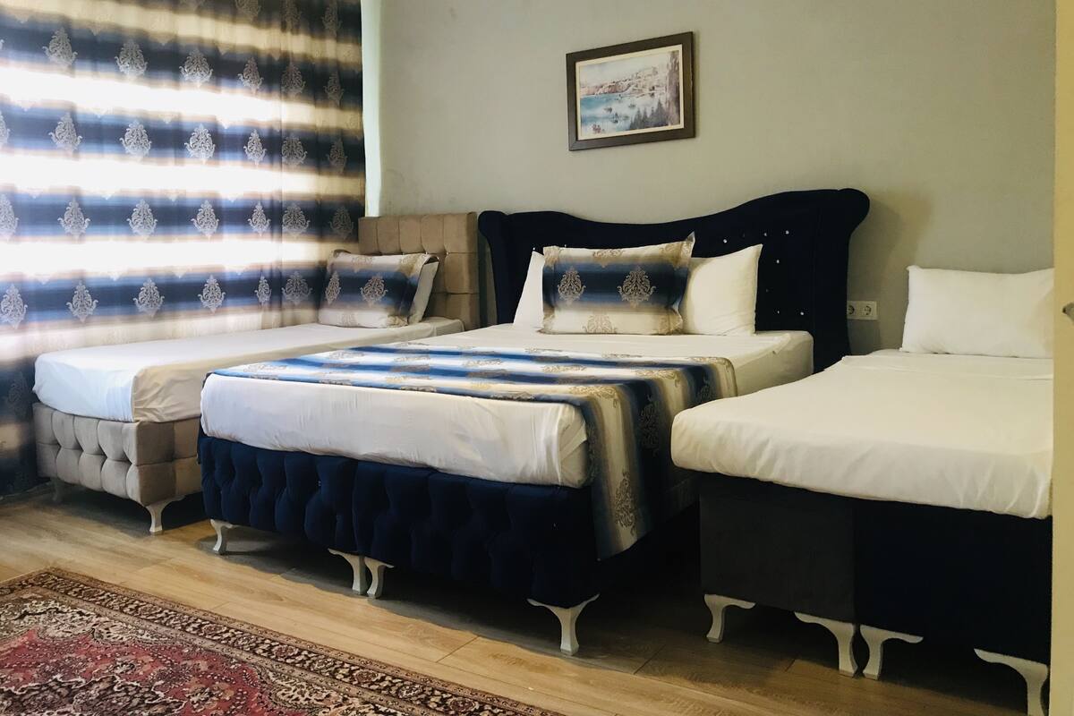 A hotel room with three beds, a large window with patterned curtains, a framed picture on the wall, and a red patterned rug on the floor.