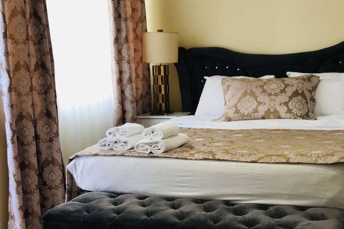 A neatly made bed with rolled towels, a black upholstered bench, and a patterned throw blanket.