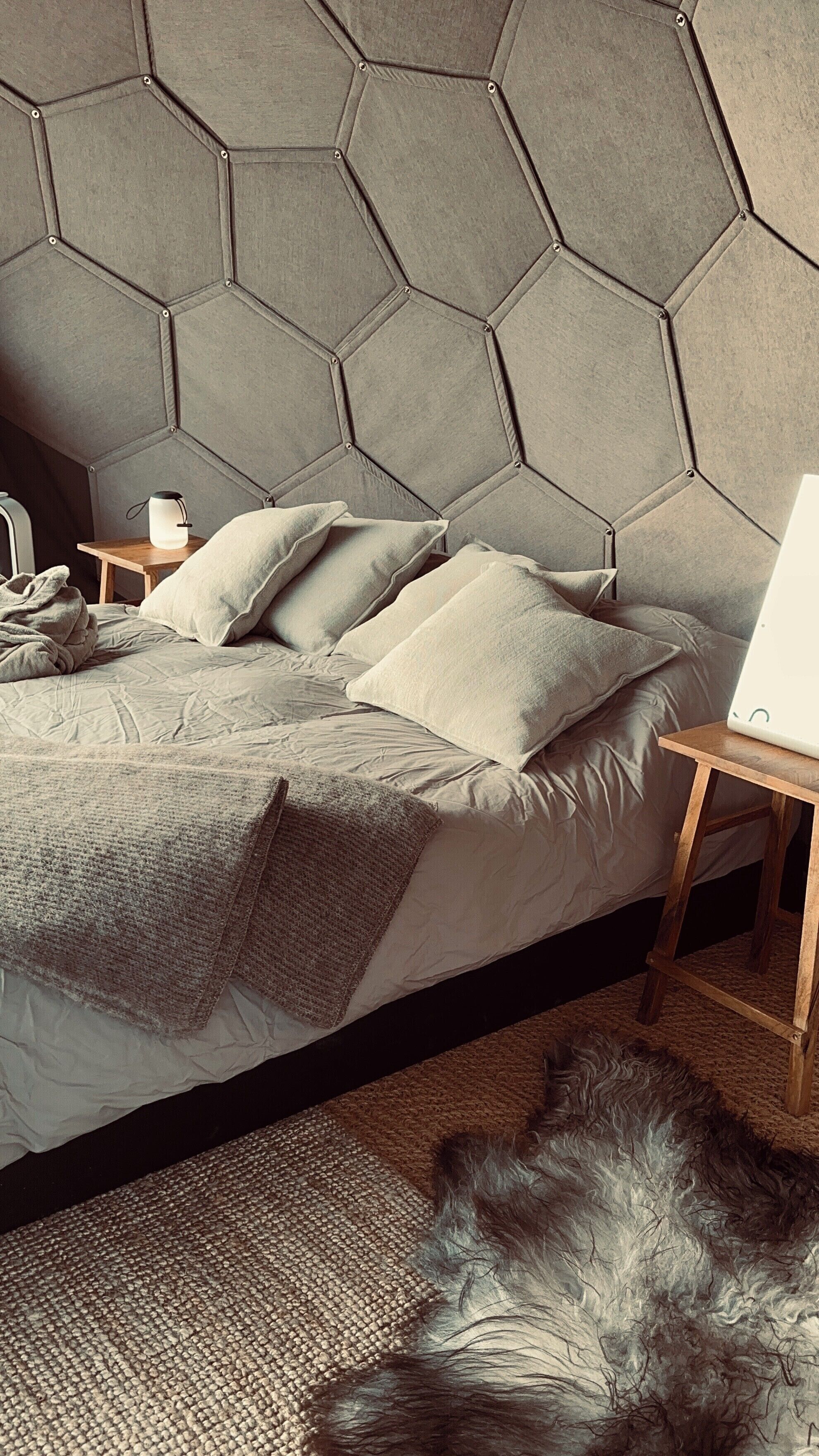 Et soveværelse med en sekskantet sengegavl, en seng med mange puder, et sengebord med en lampe og en blød måtte på gulvet.