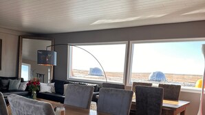 Un interior modern amb una taula de dinar de fusta, cadires tapissades de gris, una finestra gran amb vistes a un paisatge i un televisor penjat.