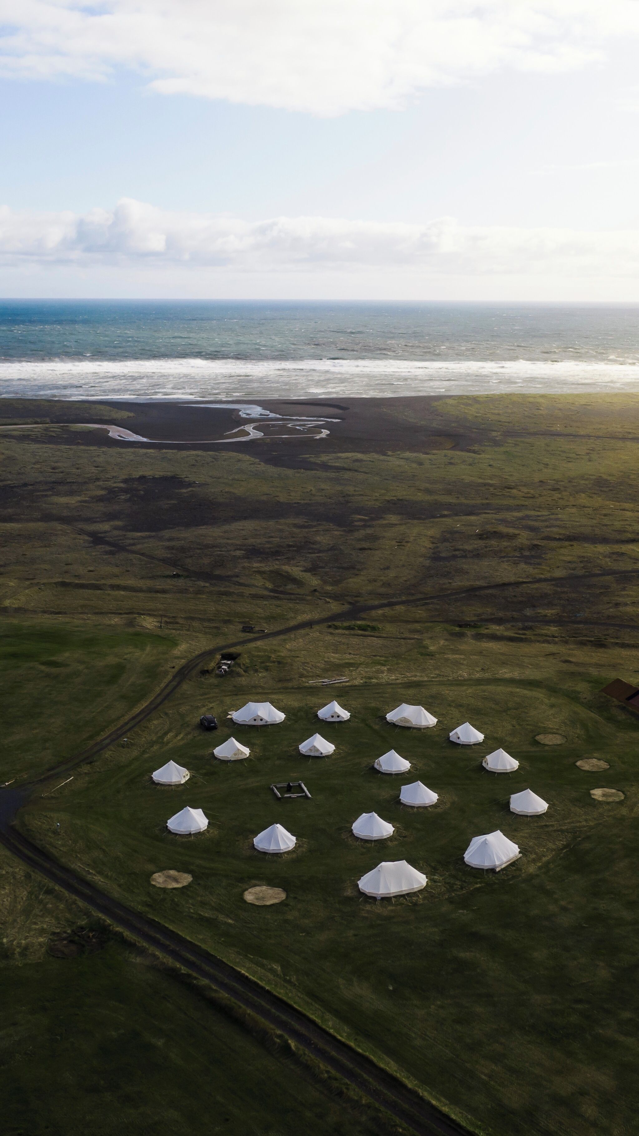 An aerial view of a campsite with white tents arranged in a semi-circle, a dirt road, and a body of water in the background.