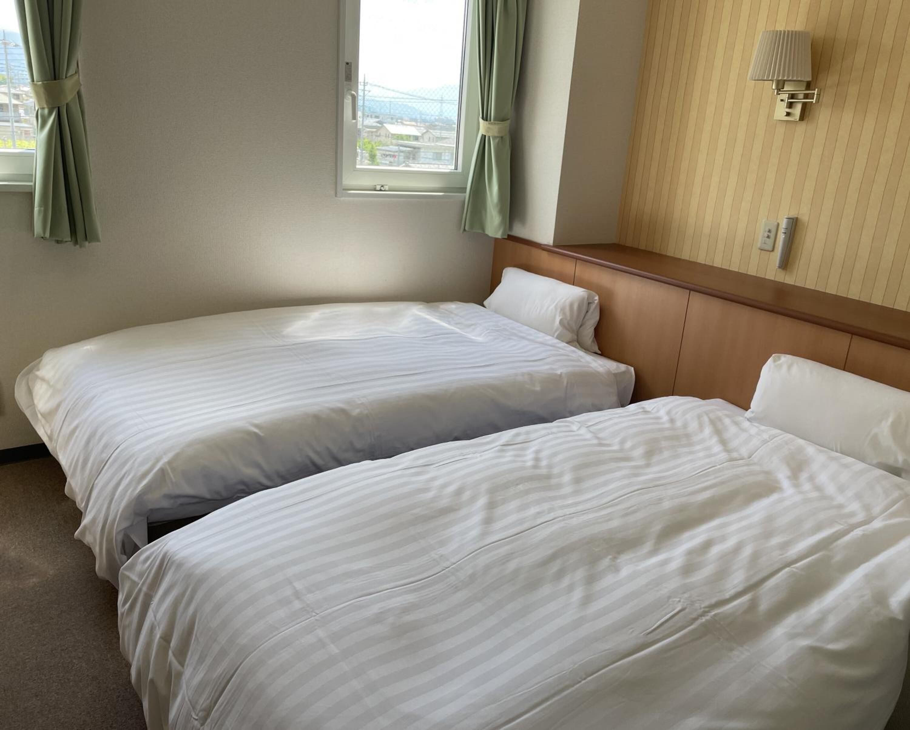 A hotel room with two beds, a window with curtains, a wall-mounted light, and a view of buildings outside.