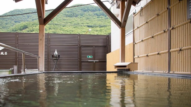 A traditional Japanese onsen with a wooden deck, a hot spring bath, and a view of a forested mountain. - Kur and Hotel Isawa (Fuefuki)