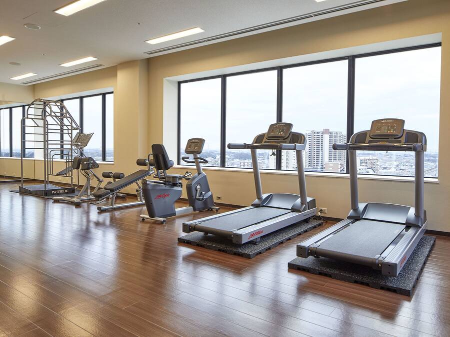A gym with treadmills, weight machines, and a stair climber. Large windows offer a city view.