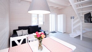 Un salon moderne avec un escalier blanc, un canapé noir, une table à manger agrémentée d’un vase de fleurs et un grand luminaire suspendu.