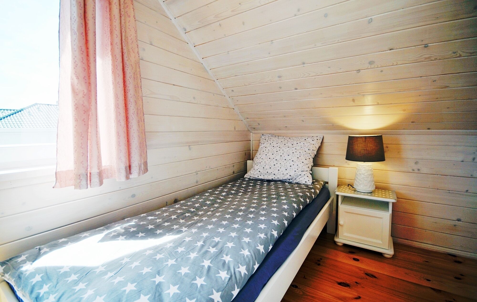 Une petite chambre avec un lit simple, une couvre-lit à motifs étoilés, une lampe de chevet et une fenêtre ornée d’un rideau rose.