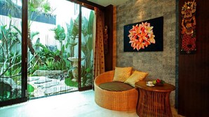 Una habitación con un gran ventanal corredero que da a un jardín tropical, un sofá de mimbre redondo con cojines, una pequeña mesita de madera y una obra de arte floral enmarcada en la pared.