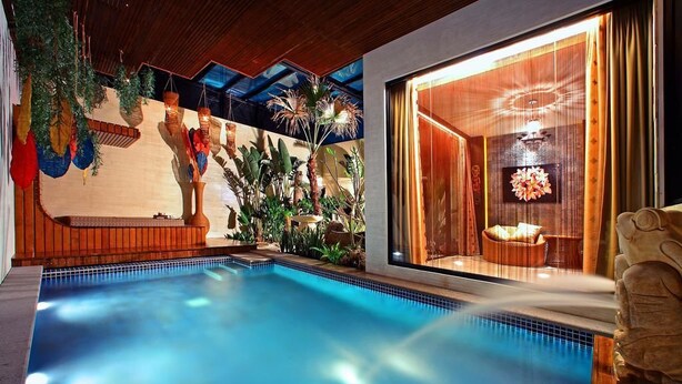 Una piscina con cubierta de madera, rodeada de plantas tropicales y un ventanal con cortinas.