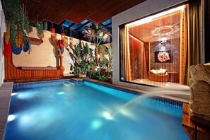Une piscine dotée d’une terrasse en bois, entourée de plantes tropicales et agrémentée d’une grande fenêtre avec des rideaux.