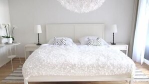 Un dormitorio con una cama blanca, dos mesitas de noche con lámparas, un candelabro blanco en el techo y una pequeña mesa blanca con una planta.