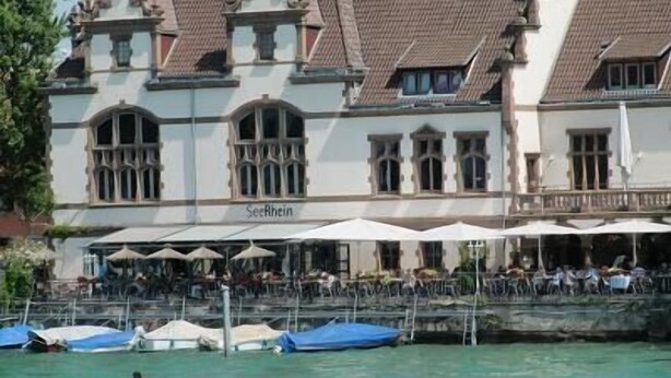 Un edificio histórico con un letrero que dice "SeeRhein" y terraza al aire libre con sombrillas.