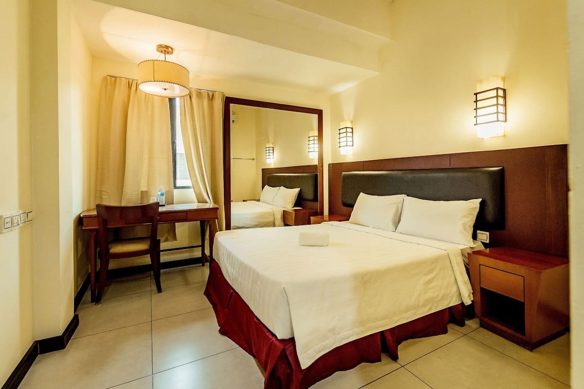 A hotel room with a bed, a desk, a chair, a nightstand, and a mirror.