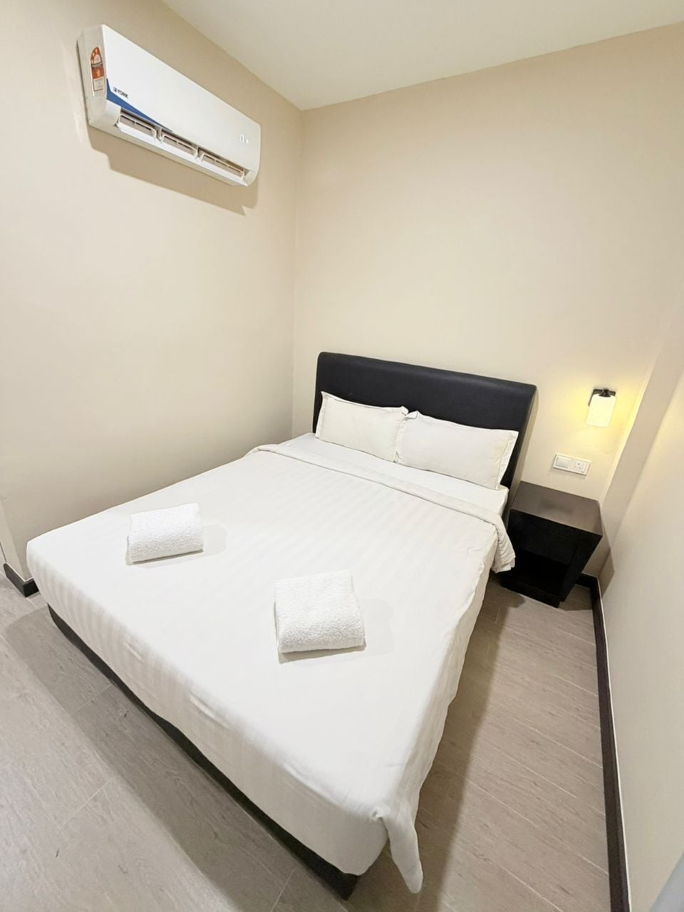 A hotel room with a bed, a nightstand, a wall-mounted air conditioner, and a wall-mounted light.