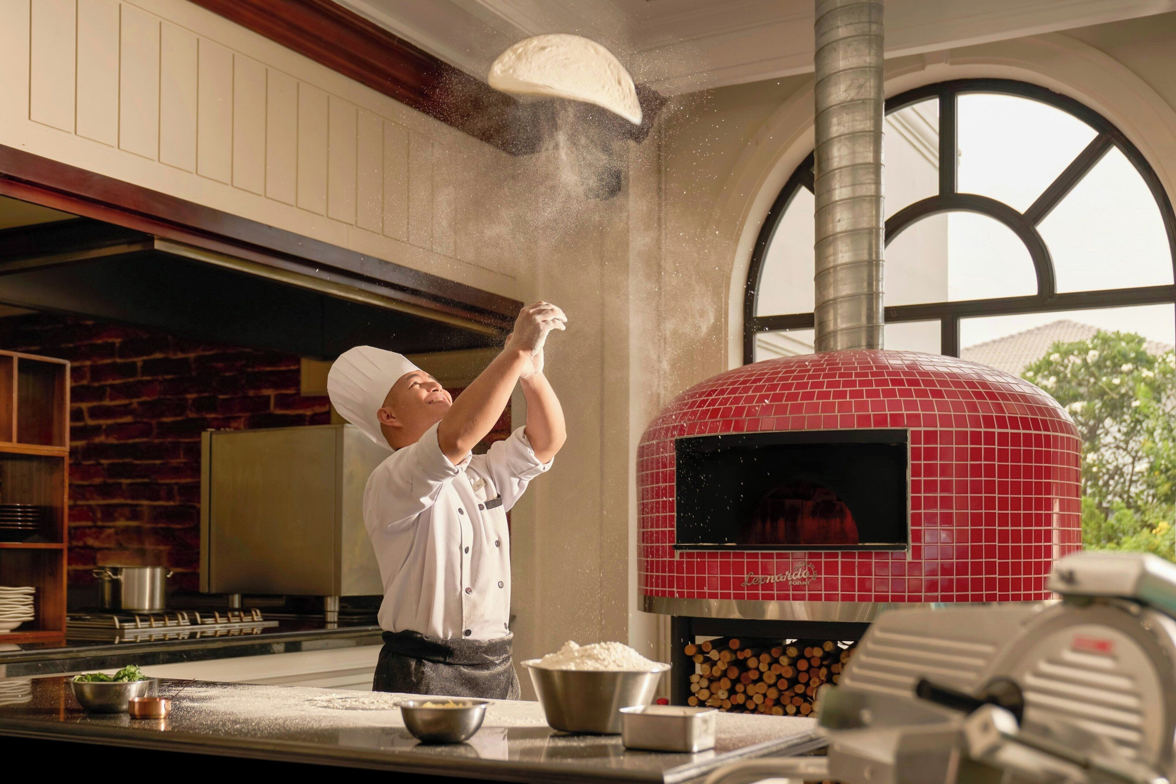 A chef tossing flour into the air in a kitchen with a red brick wall and a large window.