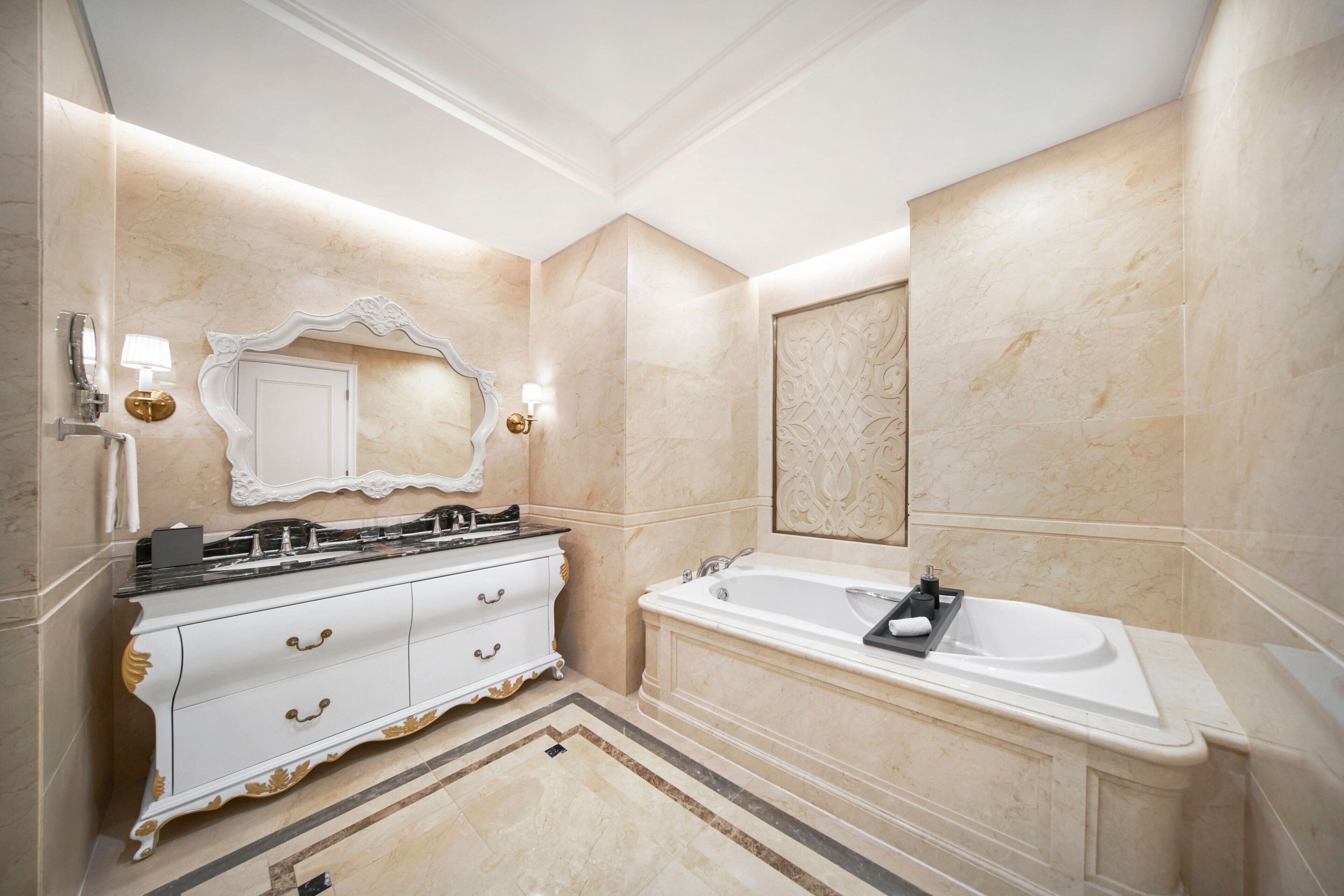 A luxurious bathroom with a large mirror, a white vanity with gold accents, a freestanding bathtub, and marble walls.