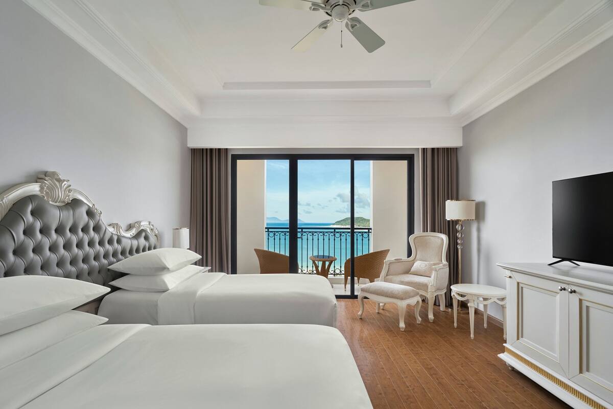 Habitación de hotel con dos camas, televisión, balcón y vista al mar, además de ventilador de techo.
