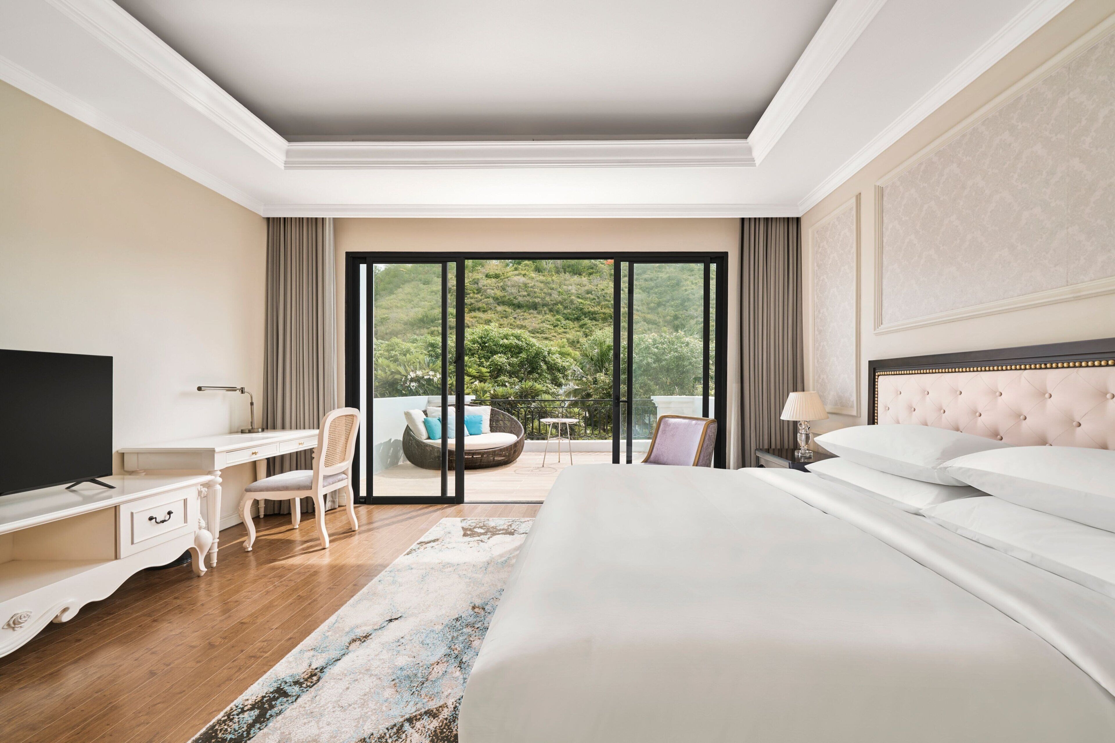 Habitación de hotel con una cama grande, un escritorio, una silla, televisor y balcón con vistas a zonas verdes.