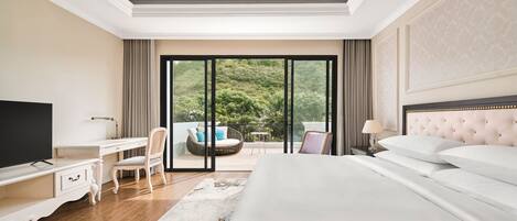 A hotel room with a large bed, a desk, a chair, a television, and a balcony with a view of greenery.