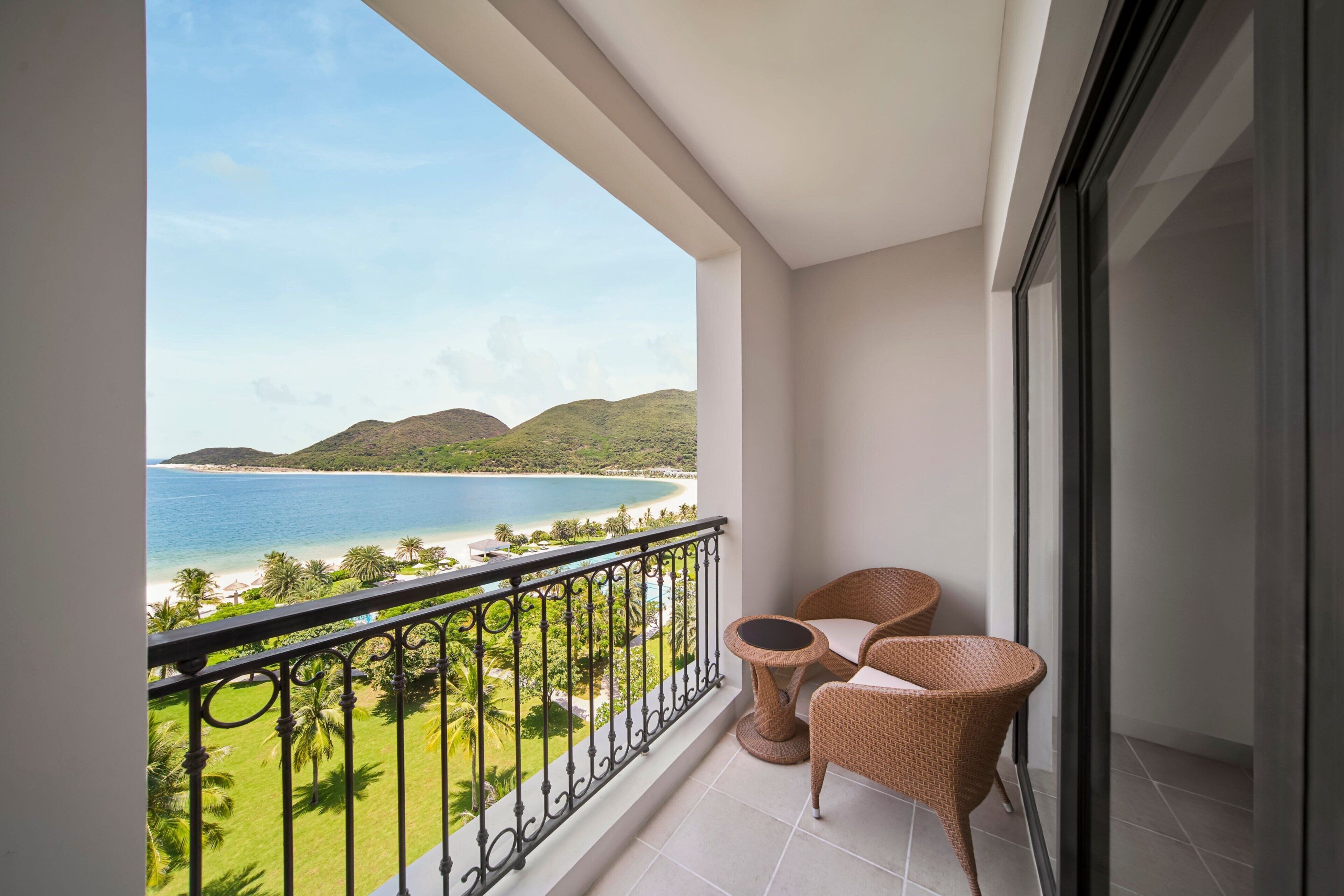 Een balkon met rieten stoelen, een glazen deur en uitzicht op een strand en bergen.