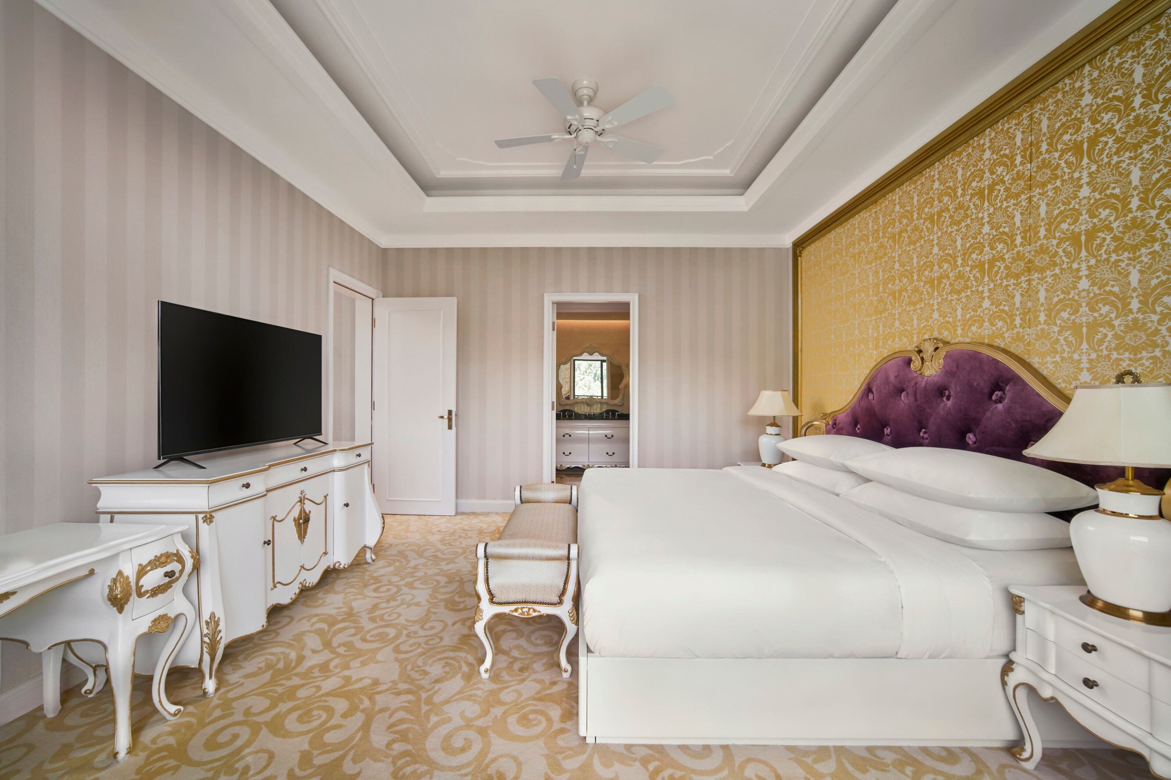 A hotel room with a large bed, a TV, a vanity with a mirror, and a ceiling fan.