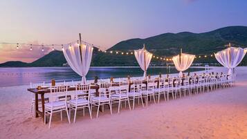 A beachside dining setup with tables and chairs, string lights, and draped fabric.