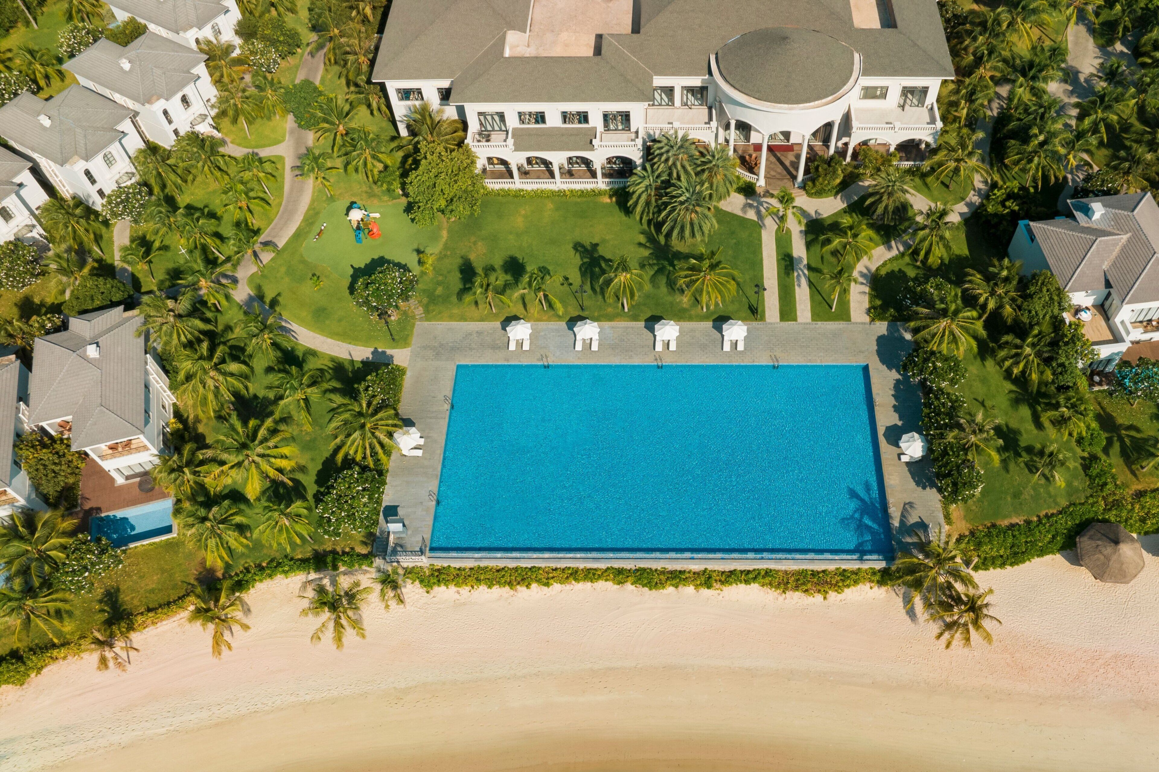 An aerial view of a resort with a large swimming pool, palm trees, and beachfront properties.