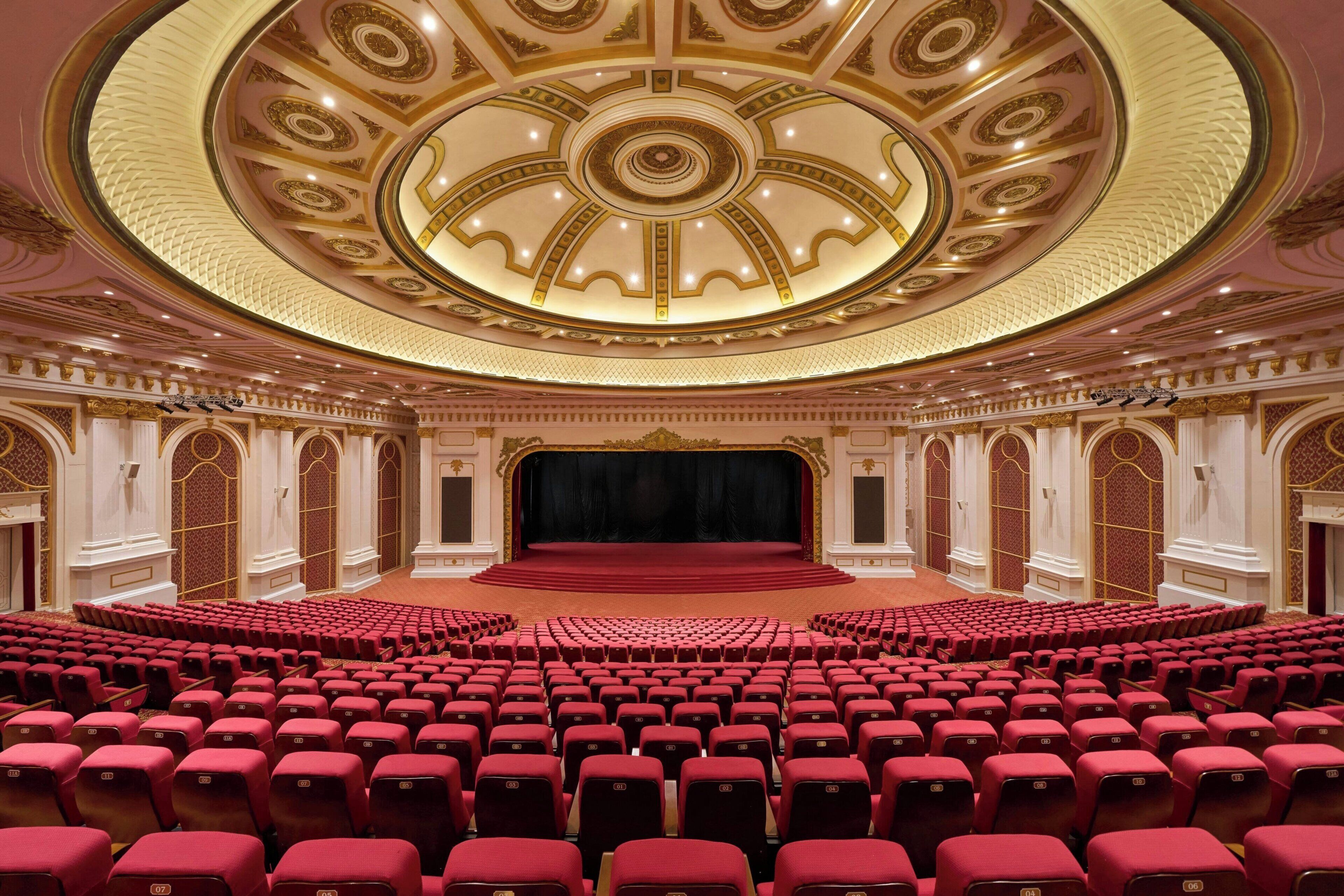 Un auditorio amplio con asientos rojos, un escenario central y decoraciones ornamentales en el techo.