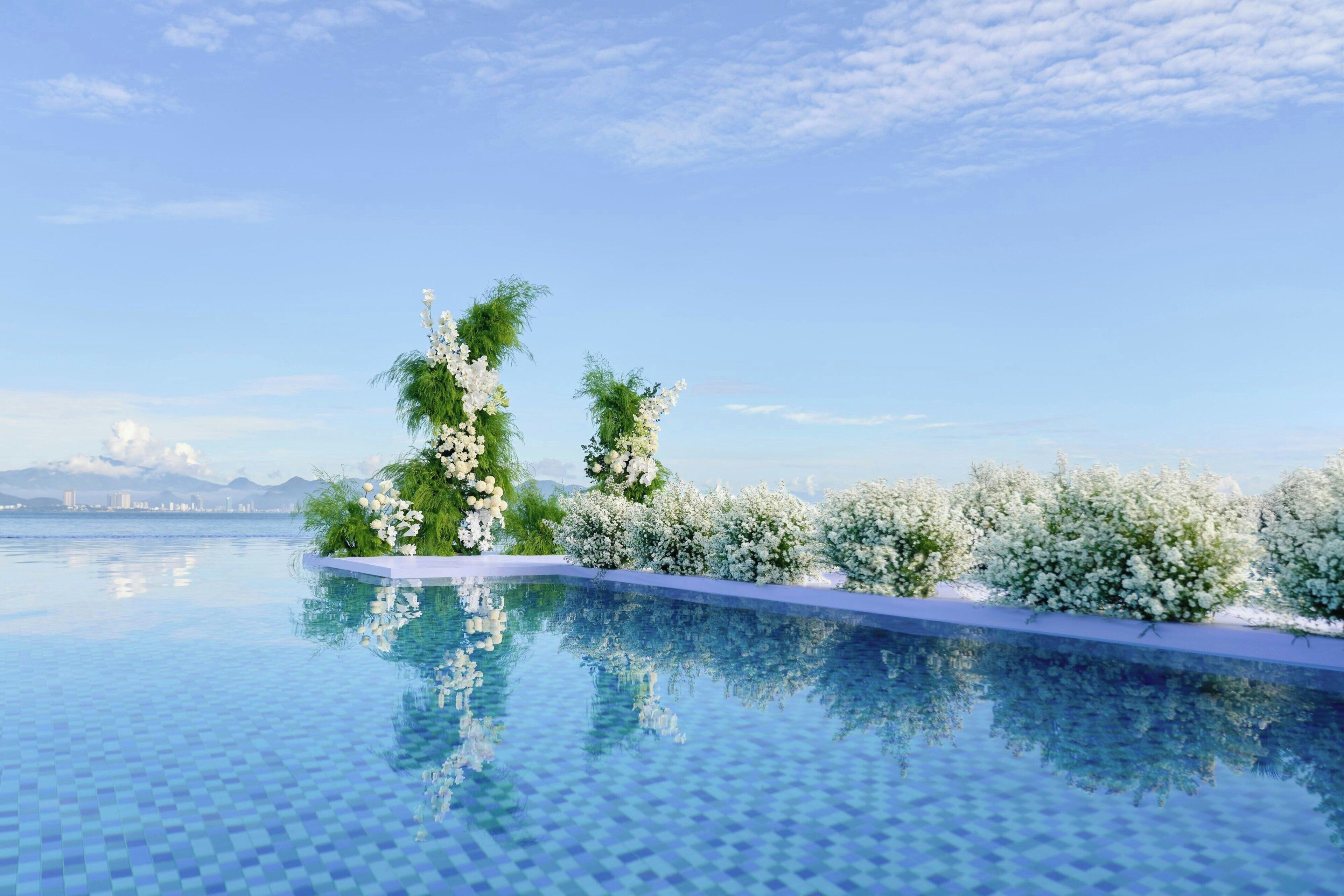 A pool with a clear blue water surface, surrounded by white flowers and greenery, with a mountain view in the background.