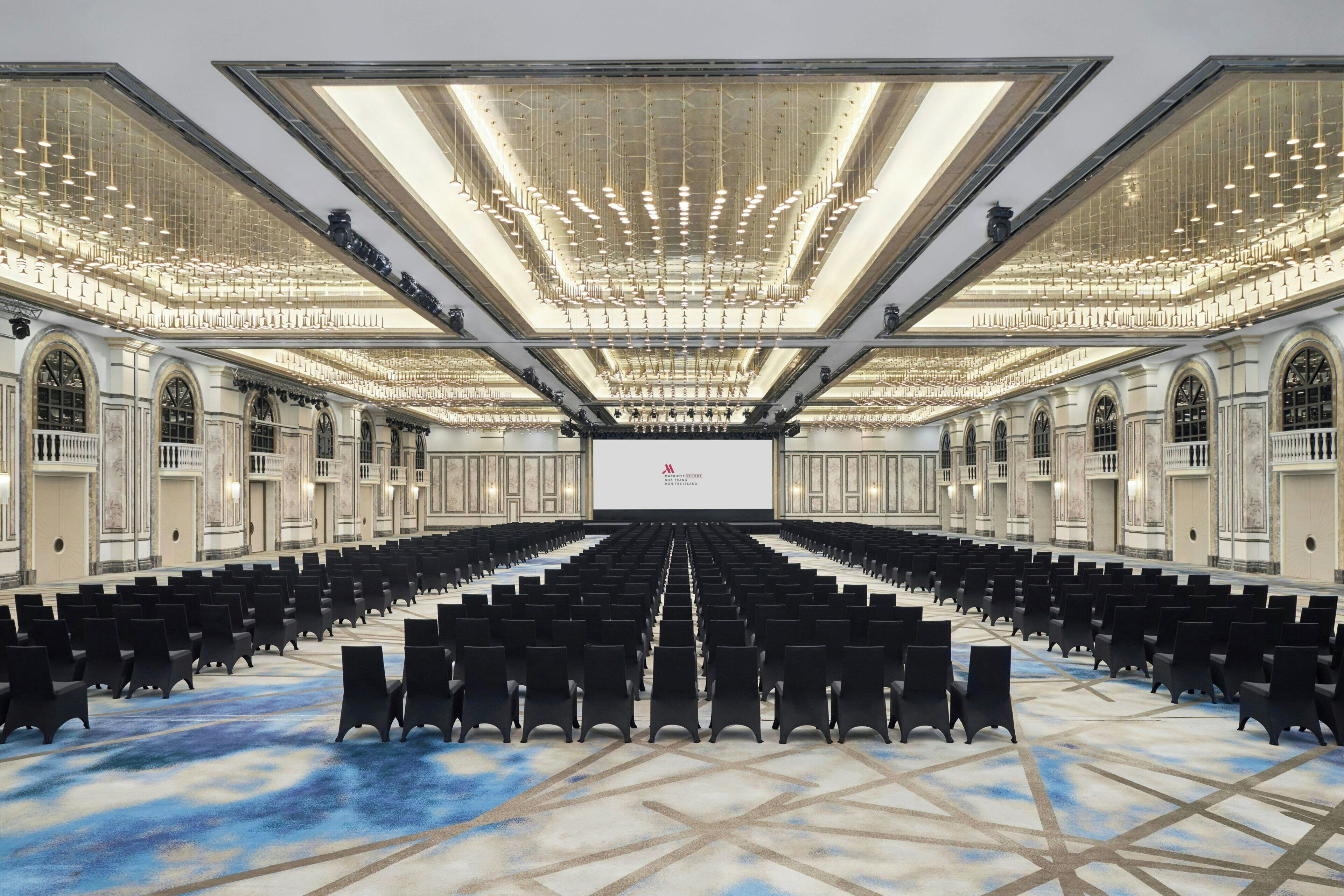 A large, well-lit conference room with rows of black chairs, a central stage, and ornate architectural details.