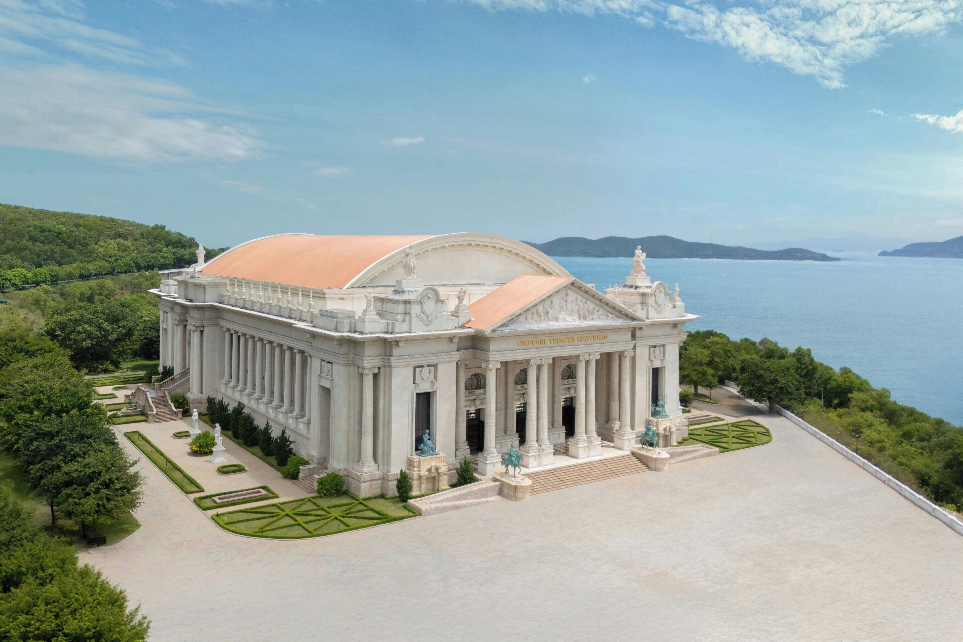 A grand building with classical architecture, featuring columns and a large dome, surrounded by well-maintained gardens and a scenic view of the sea.