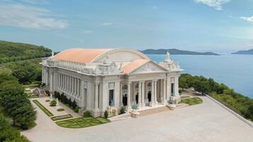 A grand building with classical architecture, featuring columns and a large dome, surrounded by well-maintained gardens and a scenic view of the sea.