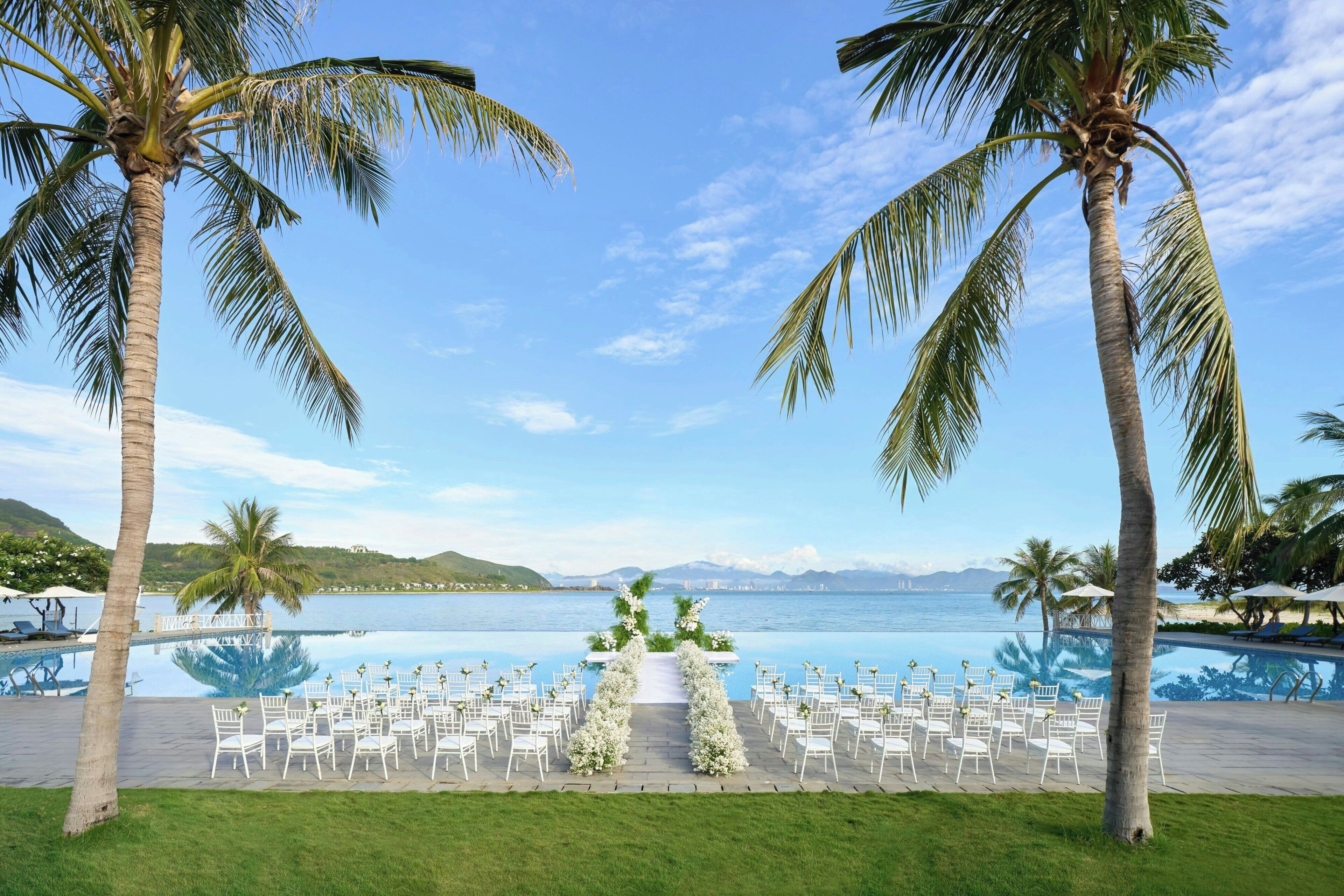 A poolside wedding venue with a clear view of the ocean, surrounded by palm trees and a well-maintained lawn.