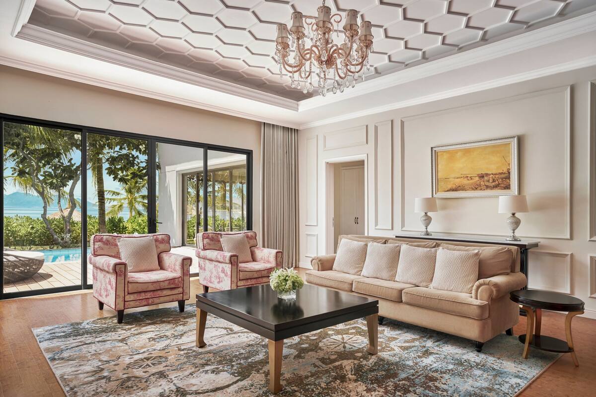 Un salón amplio con un gran ventanal, un sofá color beige, dos sillones, una mesita de centro y una araña.