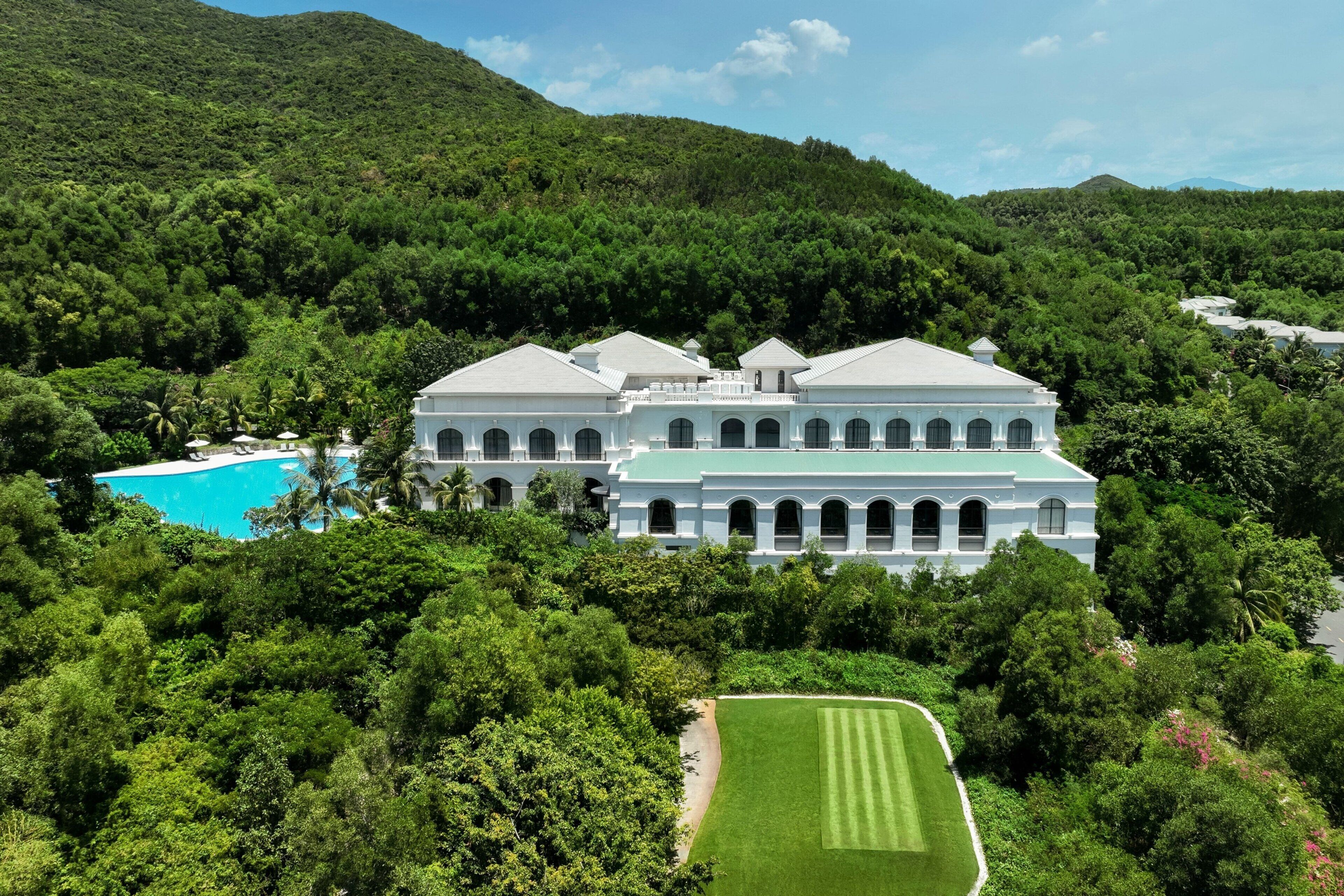 A large, white, colonial-style building surrounded by lush greenery and a pool.