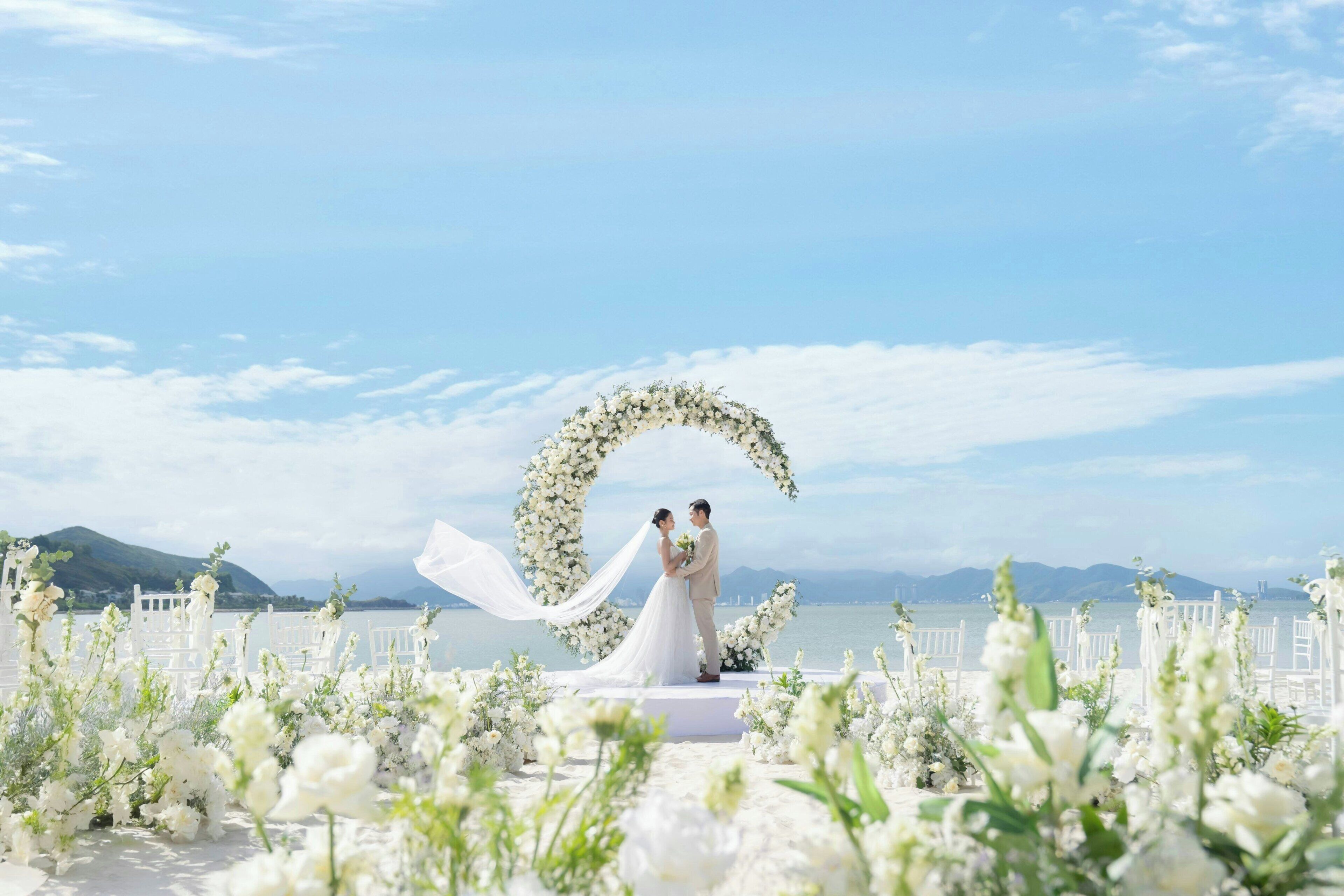 Uma cerimônia de casamento com um arco de flores e convidados sentados em cadeiras brancas.