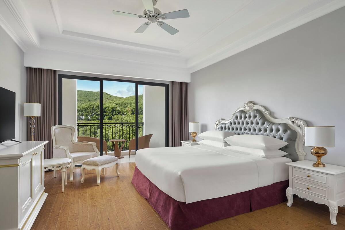 Habitación de hotel con una cama grande, un escritorio, una silla y un balcón con vistas a zonas verdes.